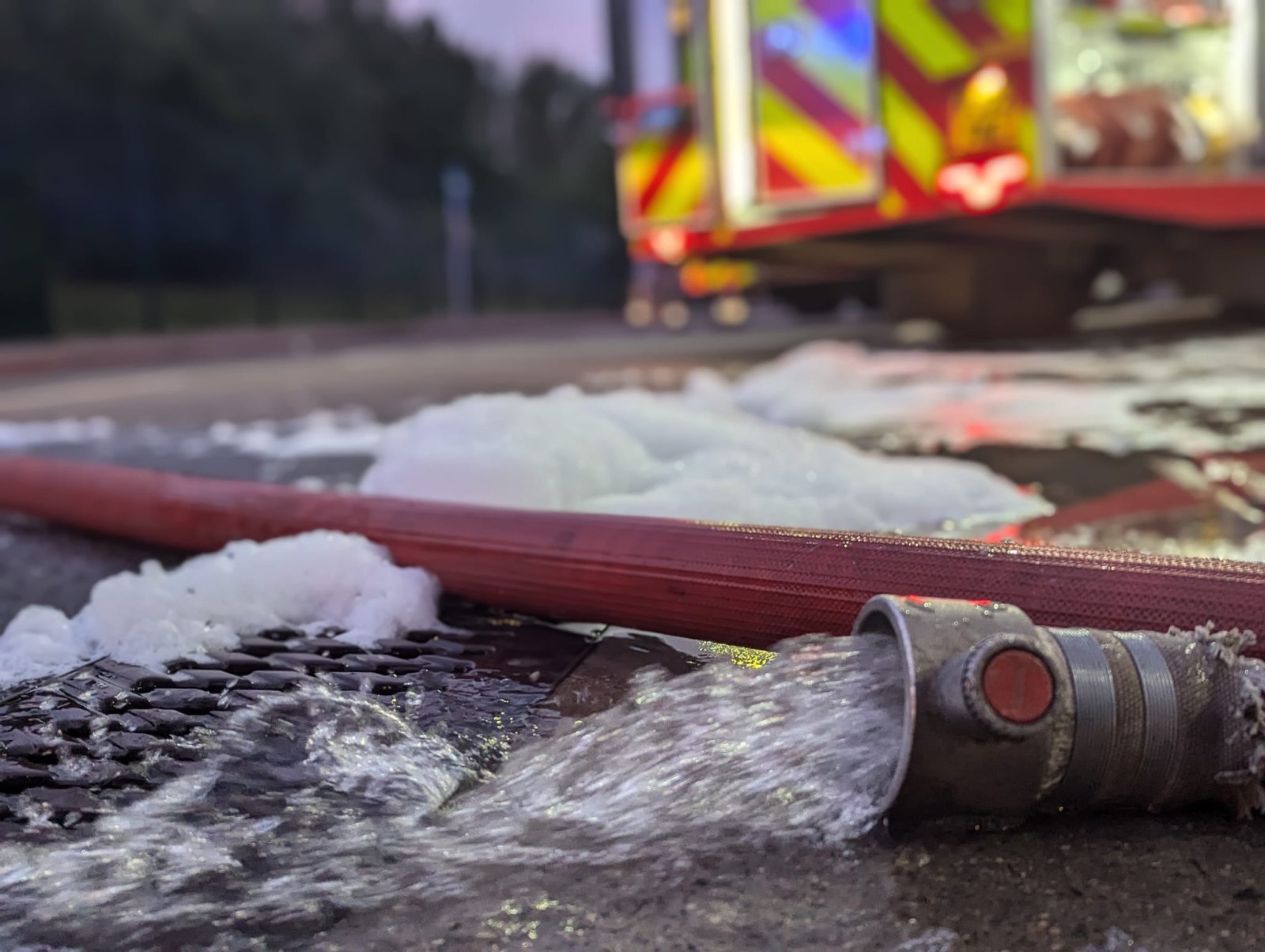 Photo of a hose laying on the ground with water coming out. A fire engine is in the background.